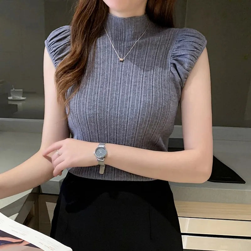 Woman wearing a gray sleeveless top with ruffled shoulders, black pants, and a watch on a blurred indoor 