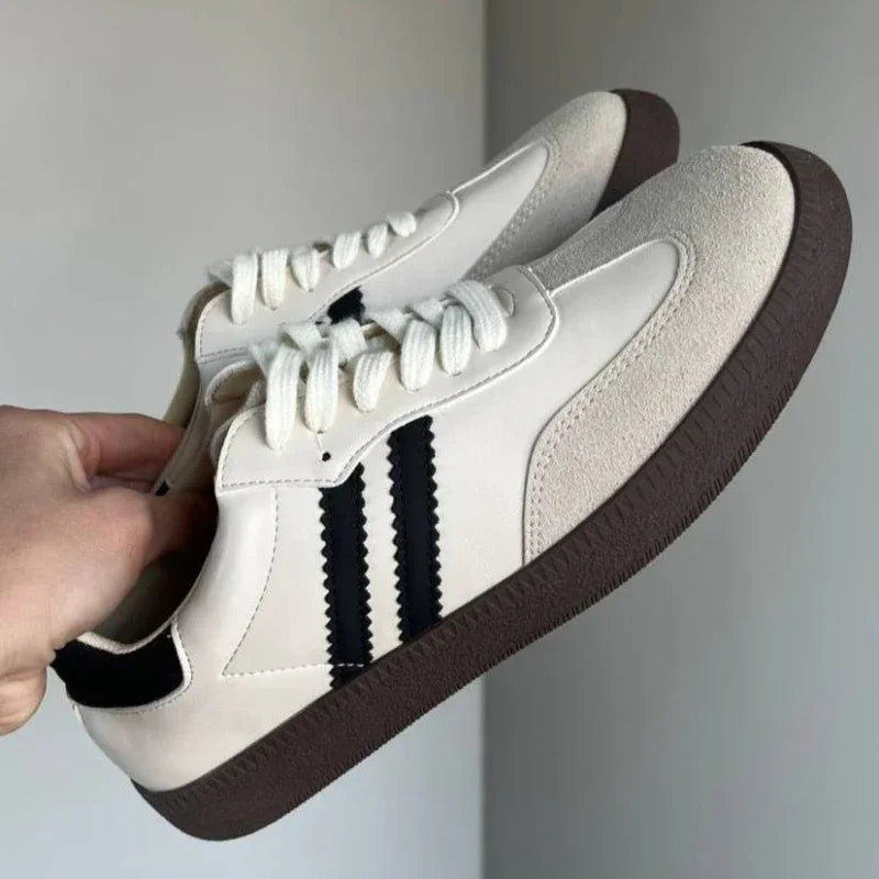 White sneakers with brown soles held by a hand against a neutral background